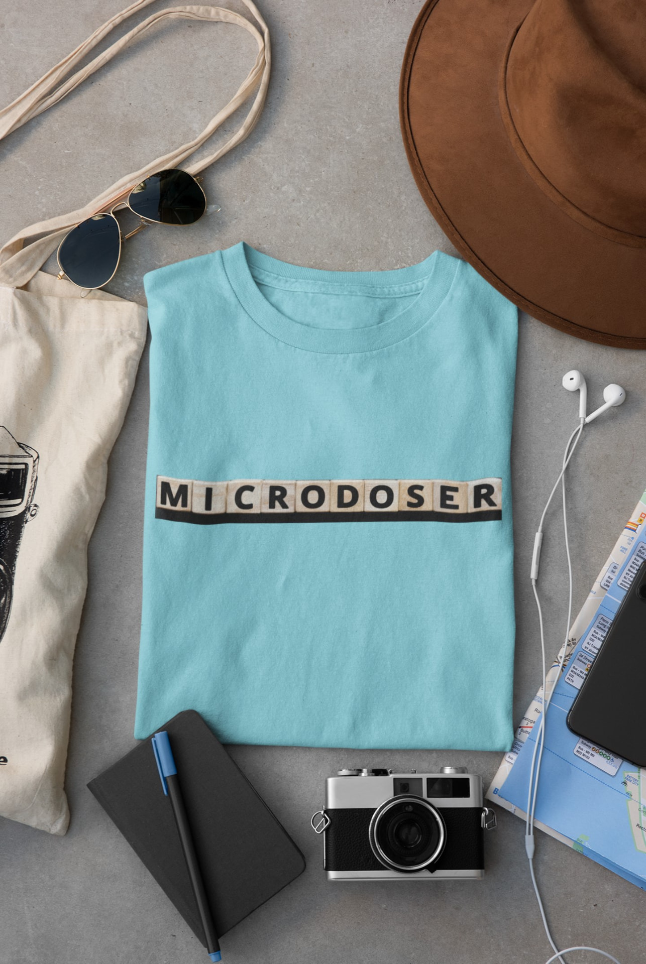 An aqua Cultivating Wisdom tee laid out with a hat, tote bag, sunglasses, and camera